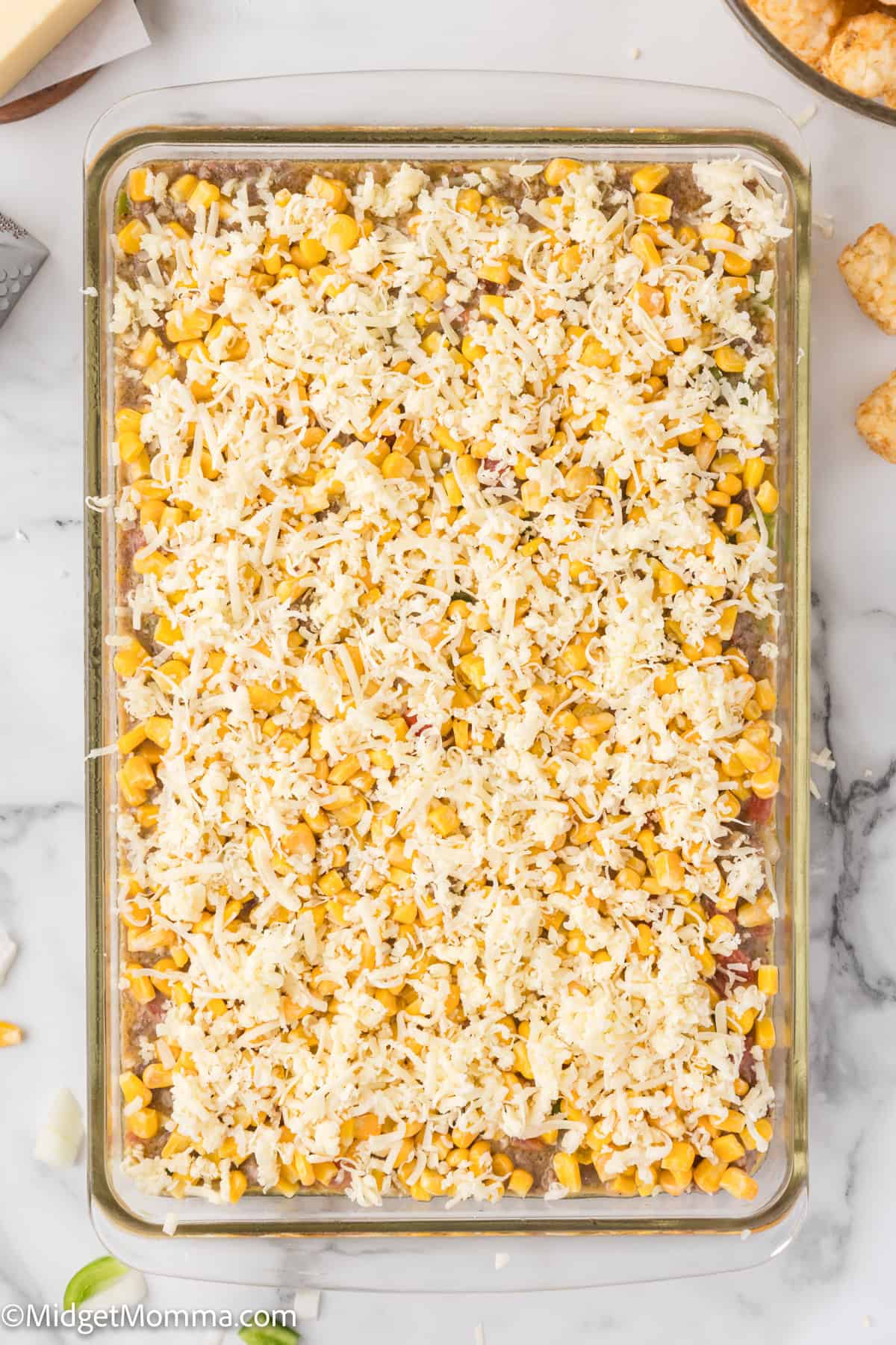 A glass baking dish filled with a casserole topped with shredded cheese and corn, resting on a marble countertop.