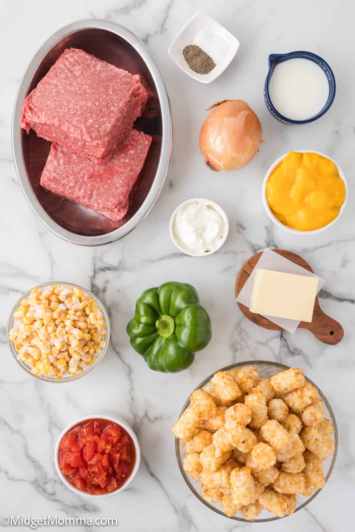 Top-down view of ingredients including ground beef, onion, pepper, milk, shredded cheese, corn, diced tomatoes, bell pepper, block of cheese, sour cream, and tater tots arranged on a marble surface.