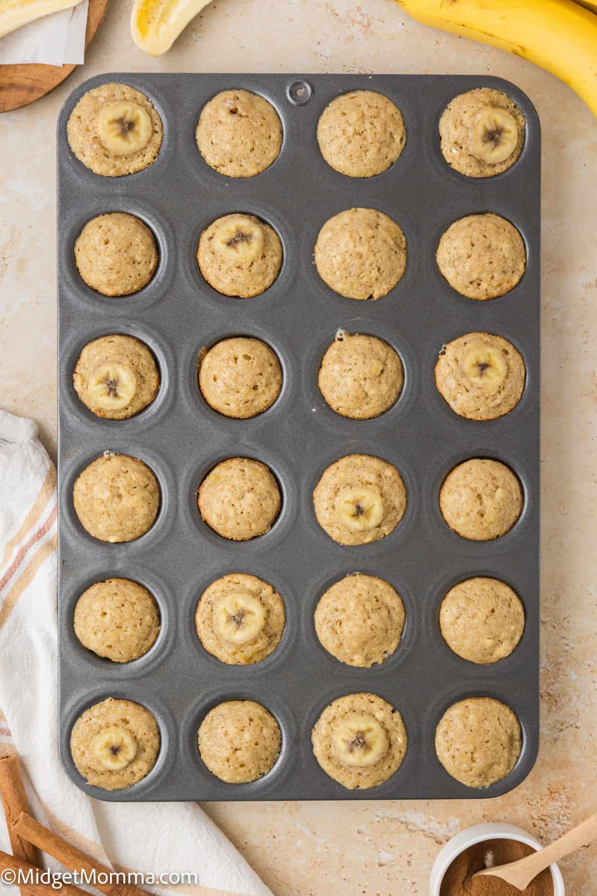 A baking tray filled with 24 mini banana muffins, some topped with banana slices, placed on a light-colored countertop with a peeled banana and a white towel nearby.