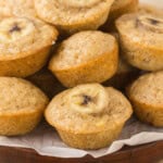 A plate of banana muffins, each topped with a banana slice, displayed on a wooden platter lined with parchment paper.