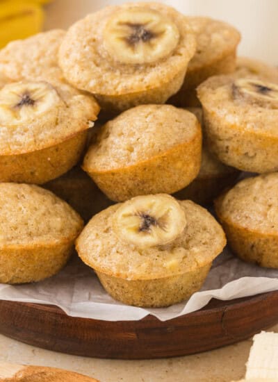 A plate of banana muffins, each topped with a banana slice, displayed on a wooden platter lined with parchment paper.