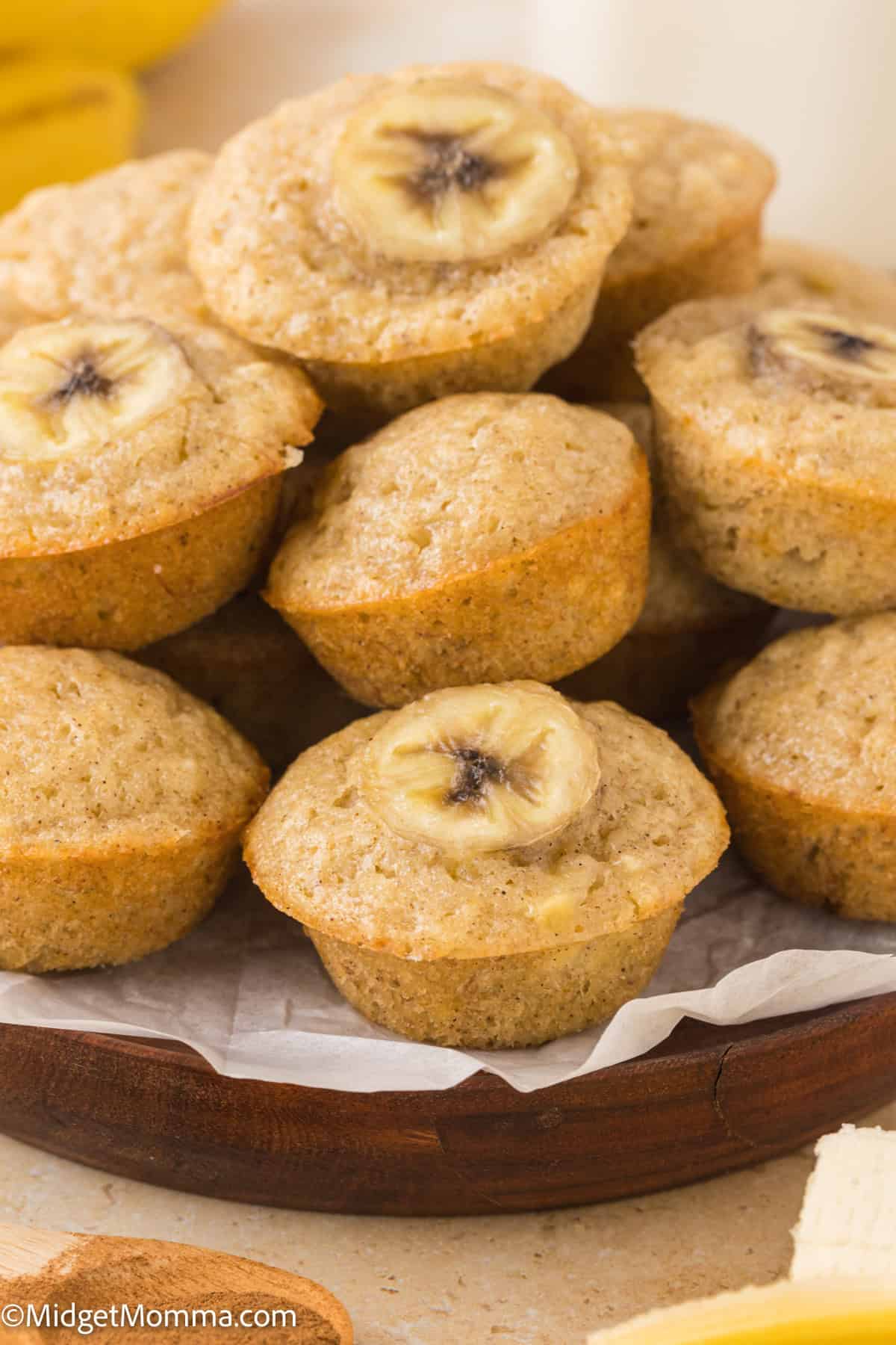 A plate of banana muffins, each topped with a banana slice, displayed on a wooden platter lined with parchment paper.