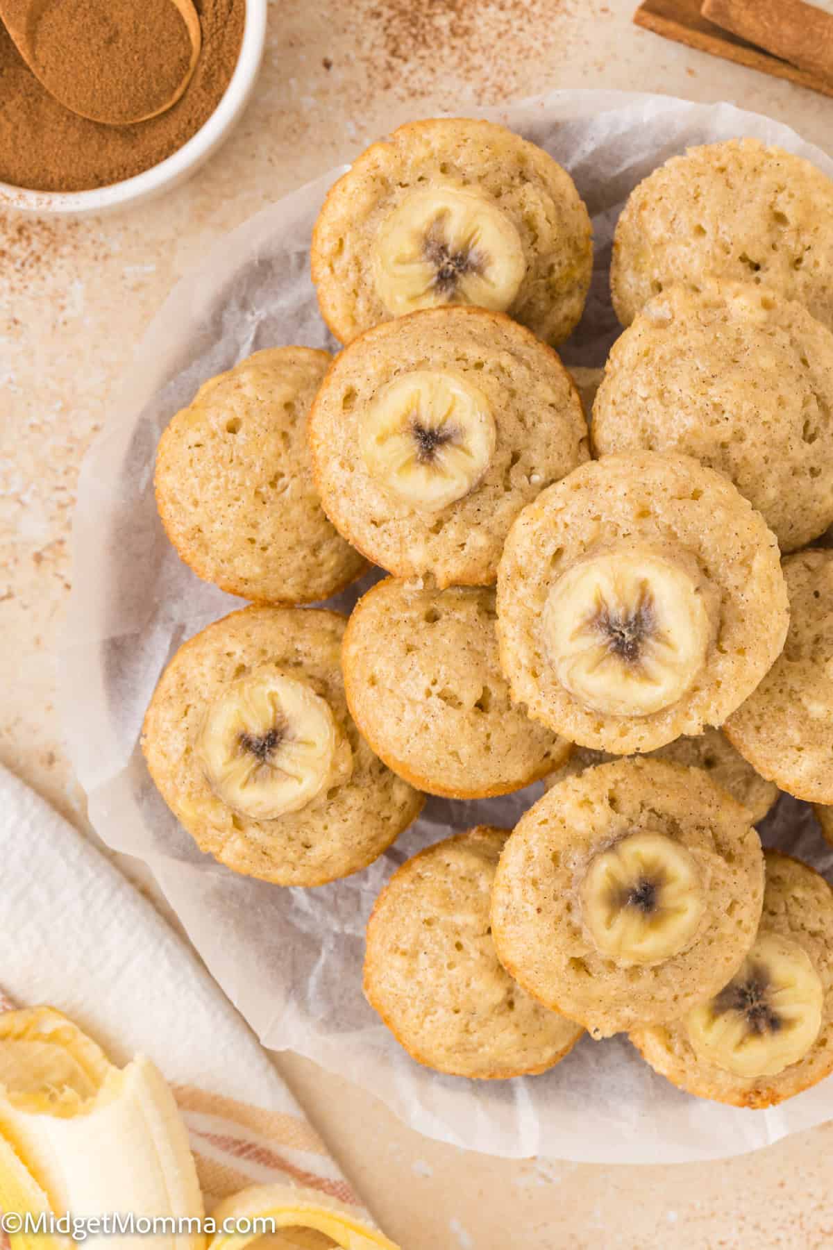 A plate of banana muffins, each topped with a banana slice. A bowl of cinnamon and banana peel segments are nearby. The background is a light-colored surface.