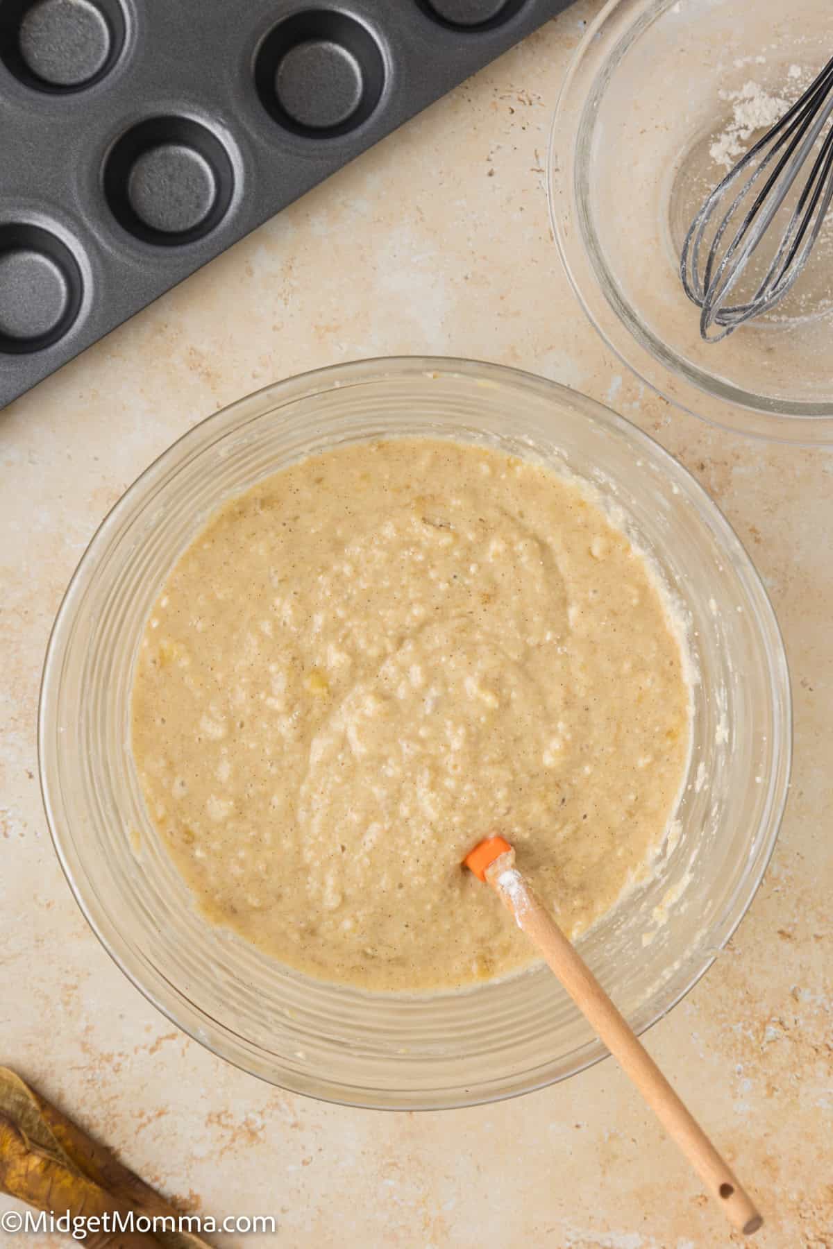 A mixing bowl contains a banana muffin batter with a wooden spoon resting in it. A whisk and empty bowl are nearby, along with a muffin tin.