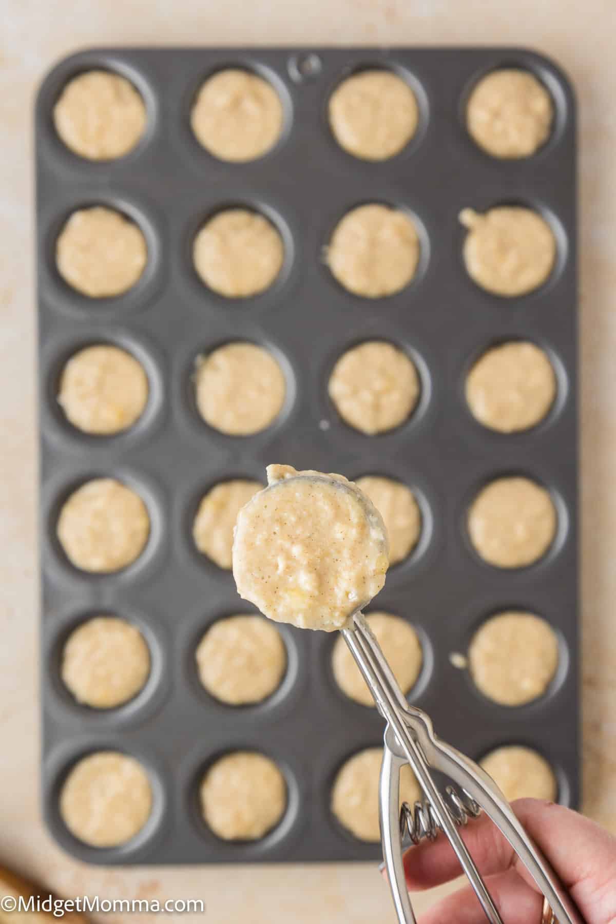 A person's hand holding a scoop of batter over a muffin tin filled with batter portions, ready for baking.