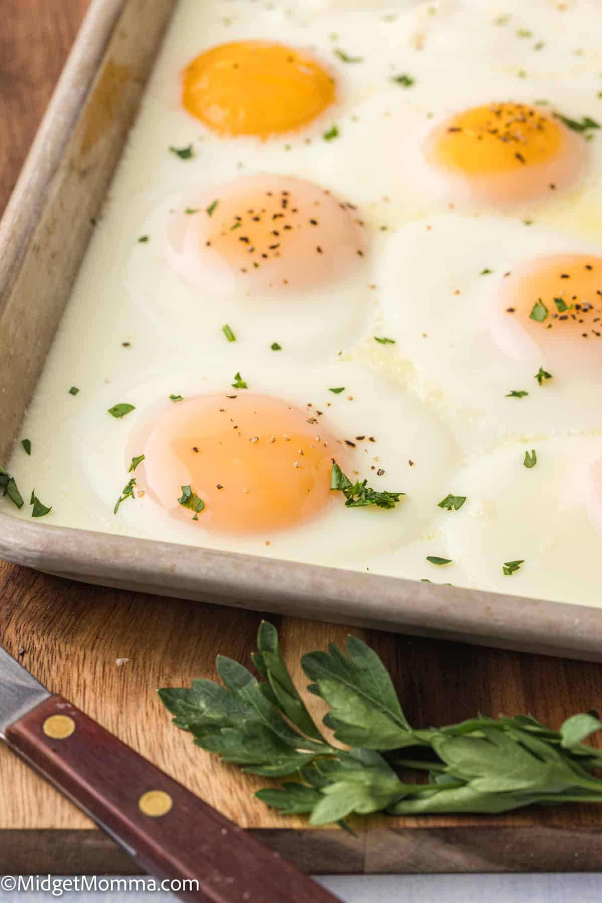 A baking sheet with six sunny-side-up eggs garnished with chopped herbs, resting on a wooden surface next to a knife and fresh herbs.