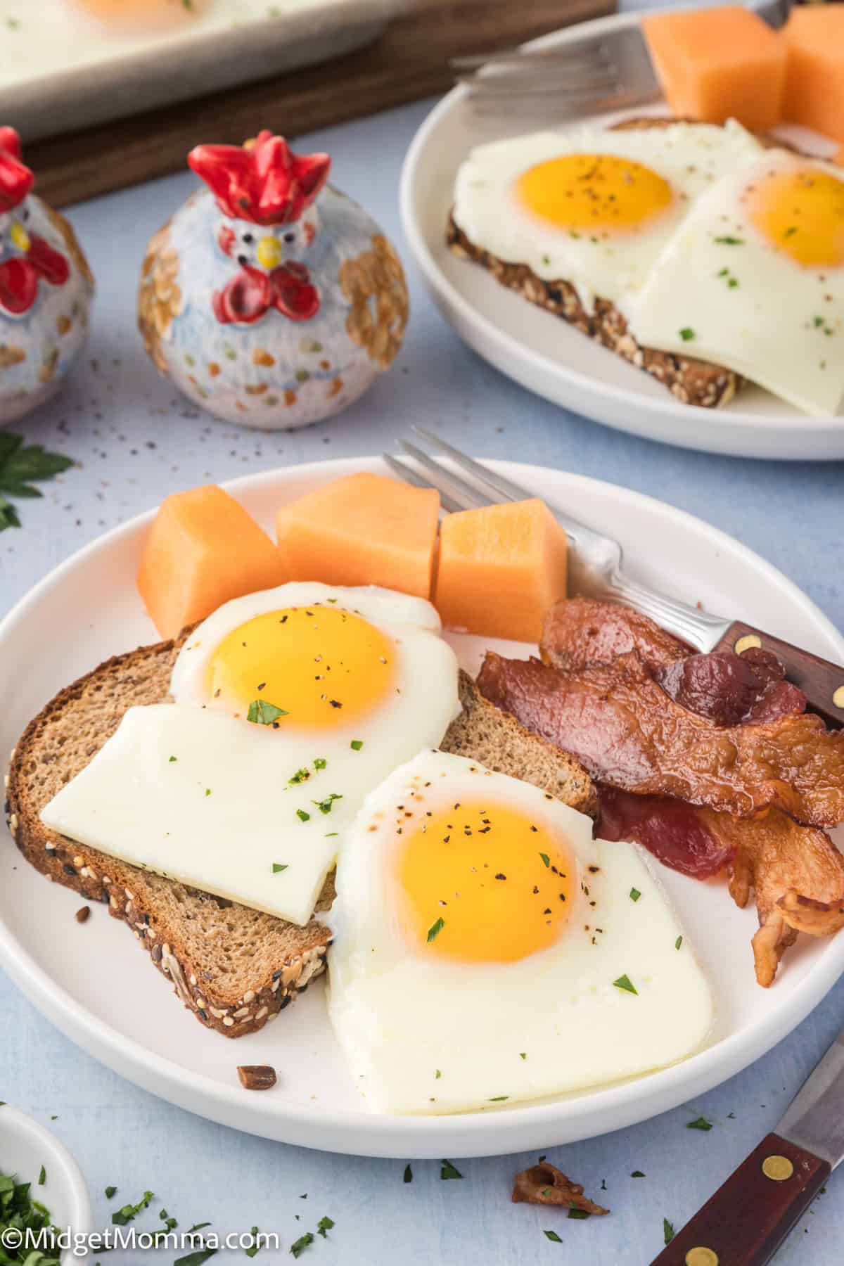 A plate with two slices of toast topped with sunny-side-up eggs, slices of cantaloupe, and crispy bacon, accompanied by salt and pepper shakers shaped like hens.