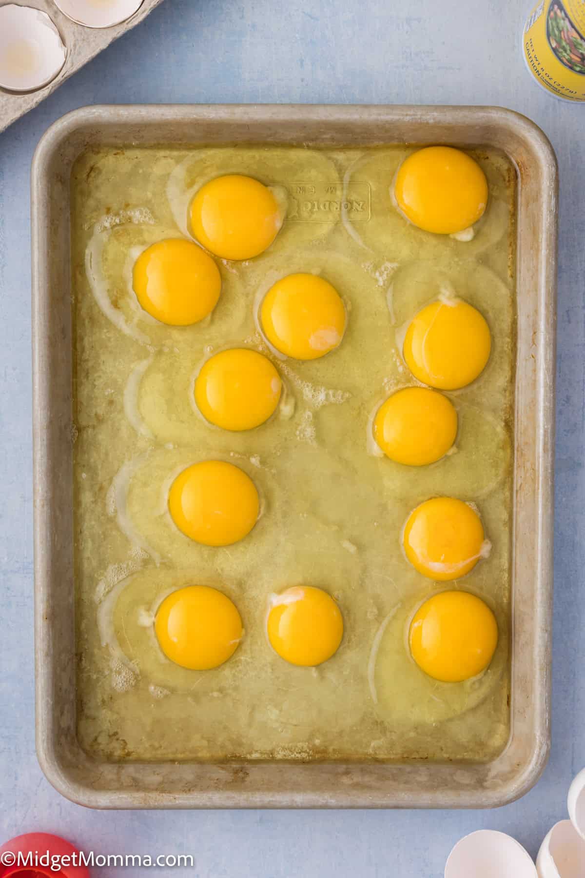 A rectangular baking pan contains a dozen raw egg yolks on top of a partially mixed egg white mixture. Eggshells are visible in the top left corner.