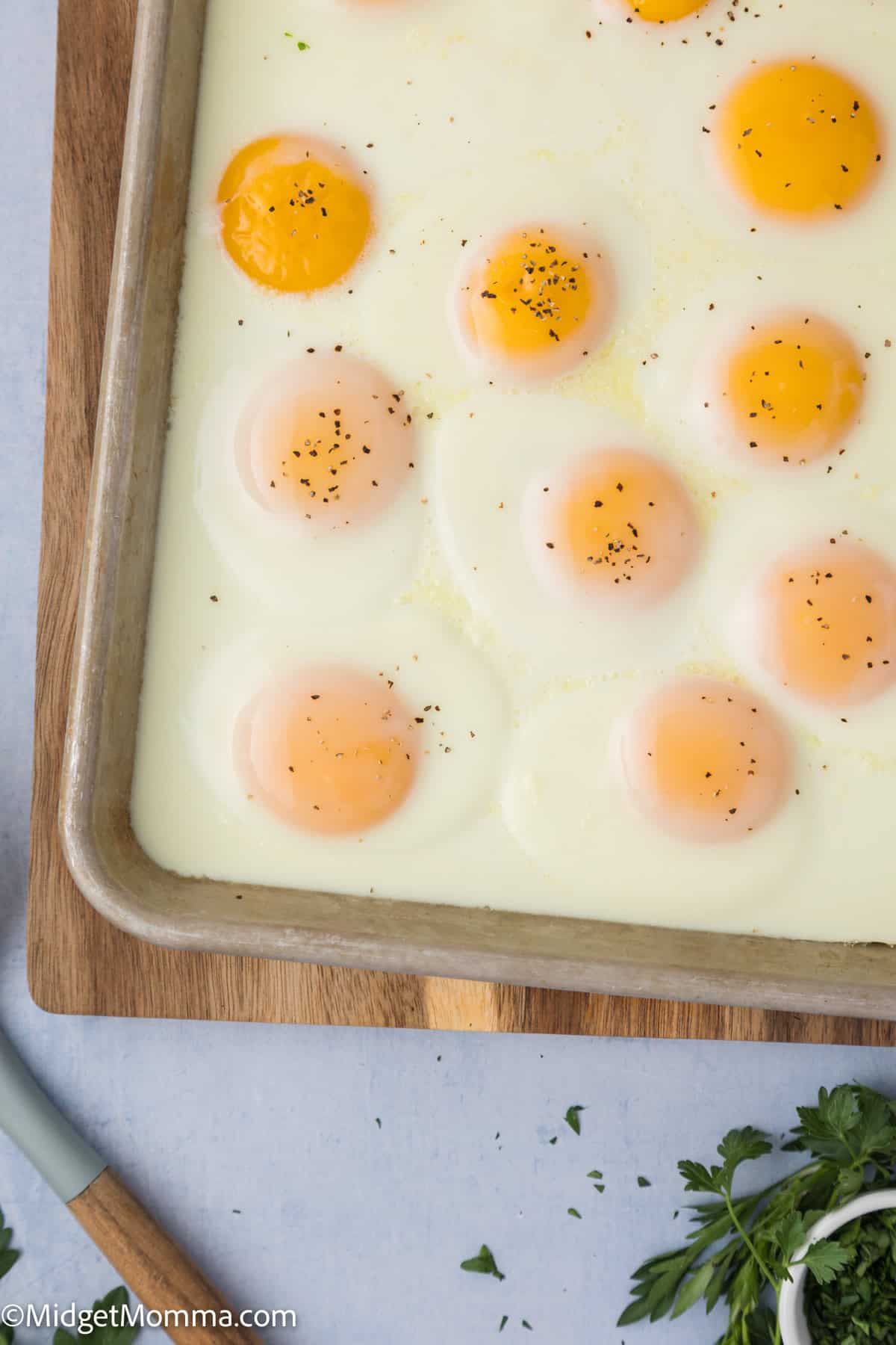 A baking sheet with eleven evenly spaced, sunny-side-up eggs sprinkled with black pepper. A knife and some fresh herbs are visible in the bottom left corner.