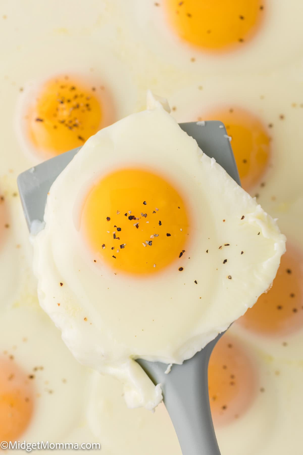 A spatula holding a cooked sunny-side-up egg with pepper, surrounded by other sunny-side-up eggs in the background.