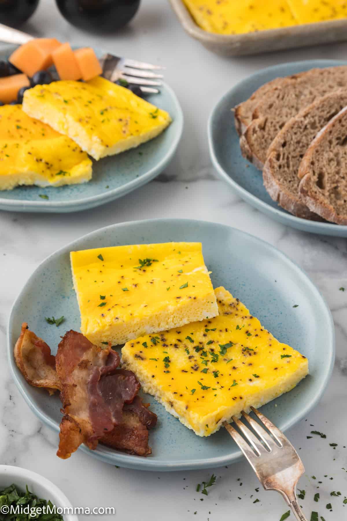 Blue plates with slices of yellow baked eggs garnished with green herbs, served with bacon slices and brown bread on the side. A fork is placed on one of the plates.