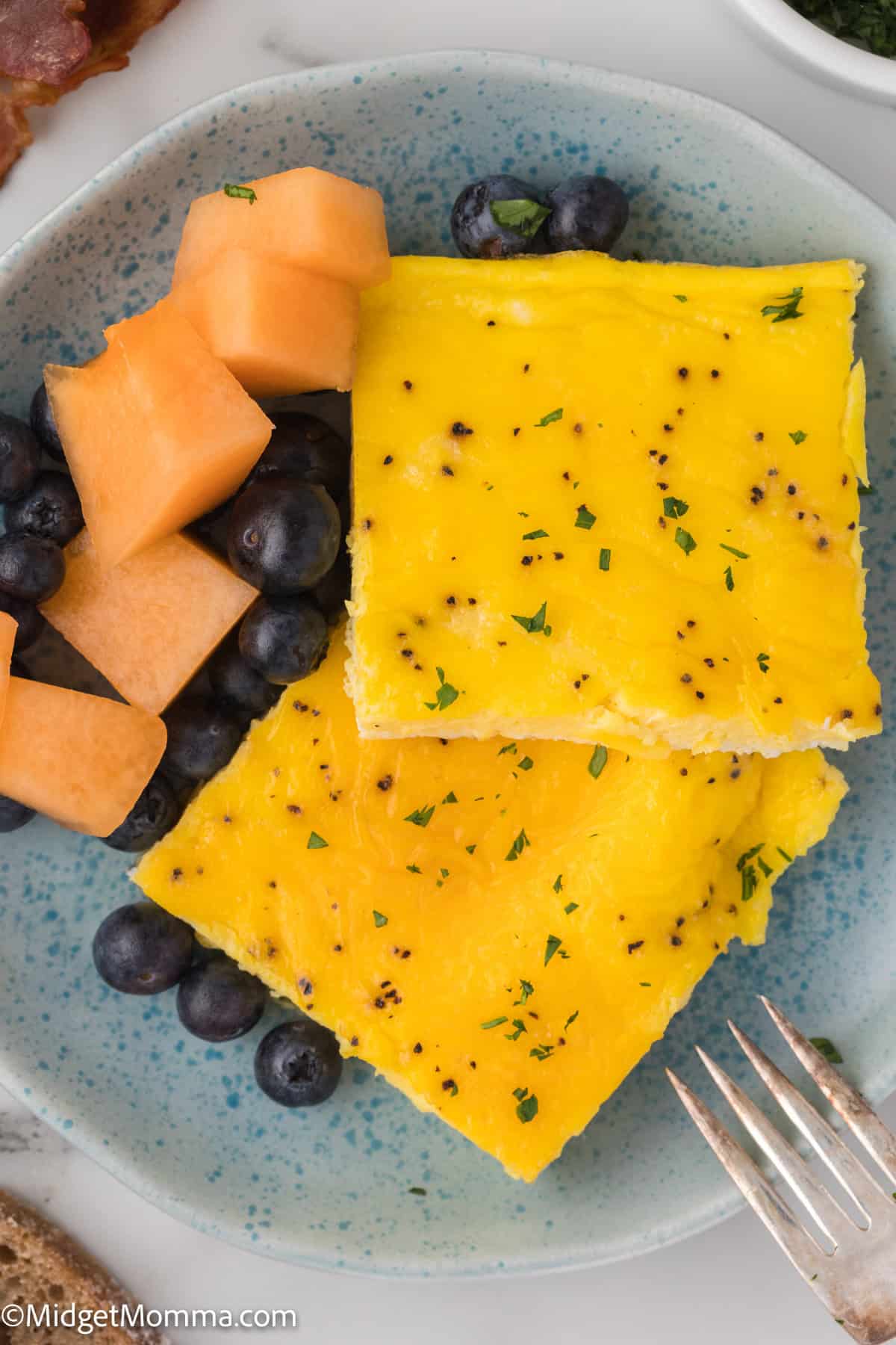 A plate with two squares of baked egg, garnished with herbs. The side includes cantaloupe and blueberries, with a fork placed near the edge of the plate.