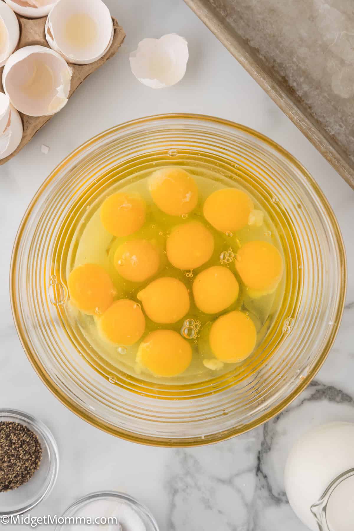 A glass bowl containing twelve raw eggs with broken eggshells, a tray, and ingredients such as milk and pepper on a marble countertop.