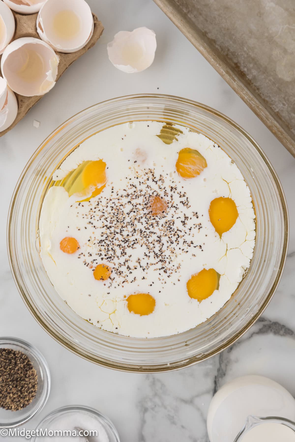 A mixing bowl with cracked eggs, milk, and black pepper on a marble countertop. Surrounding it are empty eggshells and a small container of black pepper.