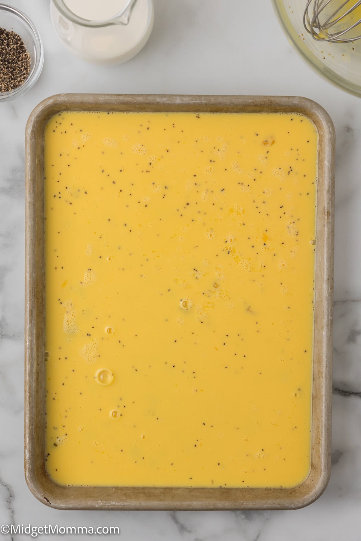 A rectangular baking dish filled with uncooked egg mixture, seasoned with black pepper. A glass jug of milk, pepper grinder, and whisk are nearby on a marble surface.