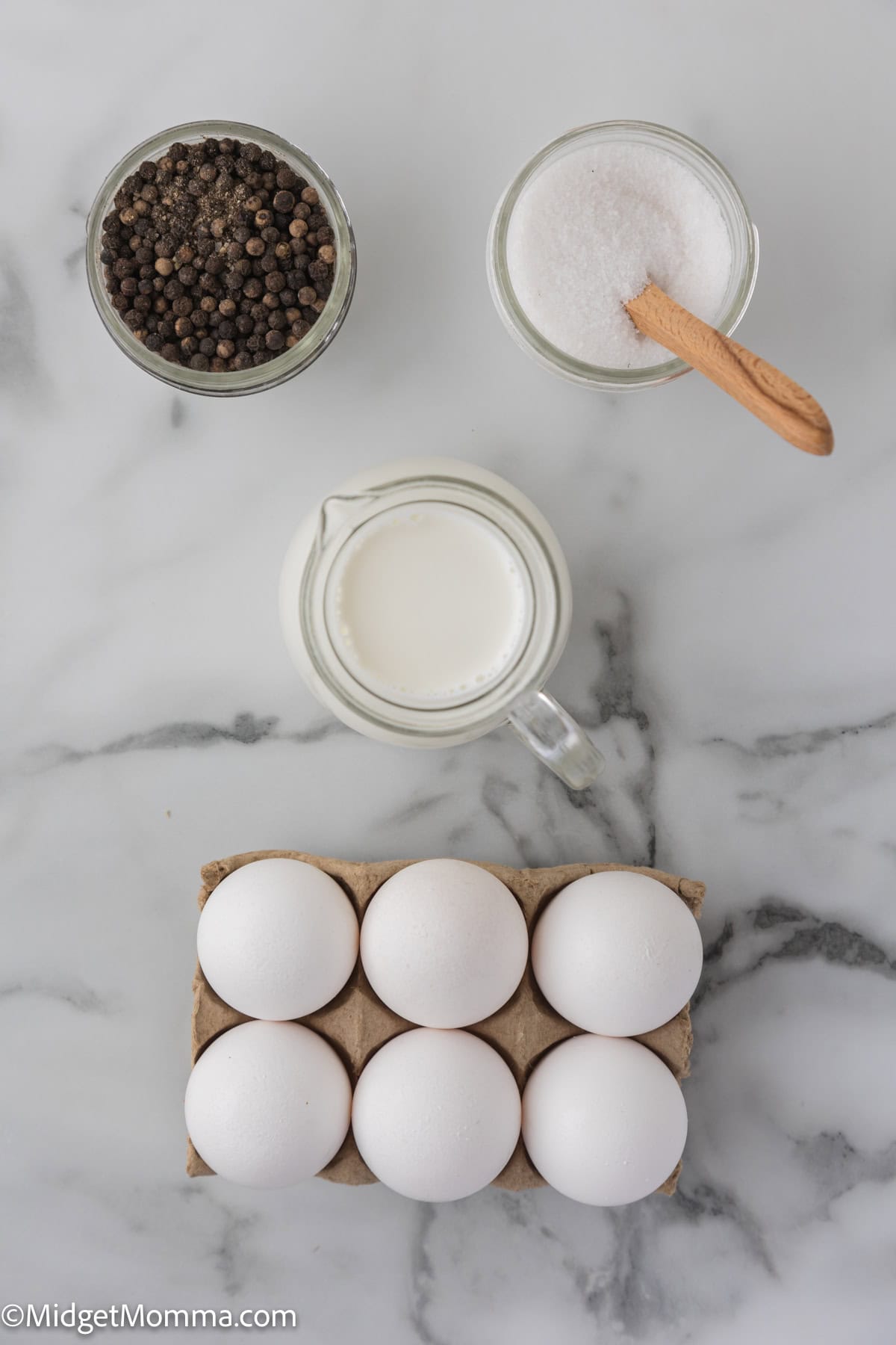 A carton with six white eggs, a glass jar of black pepper, a glass jar of salt with a spoon, and a small pitcher of milk are placed on a marble countertop.