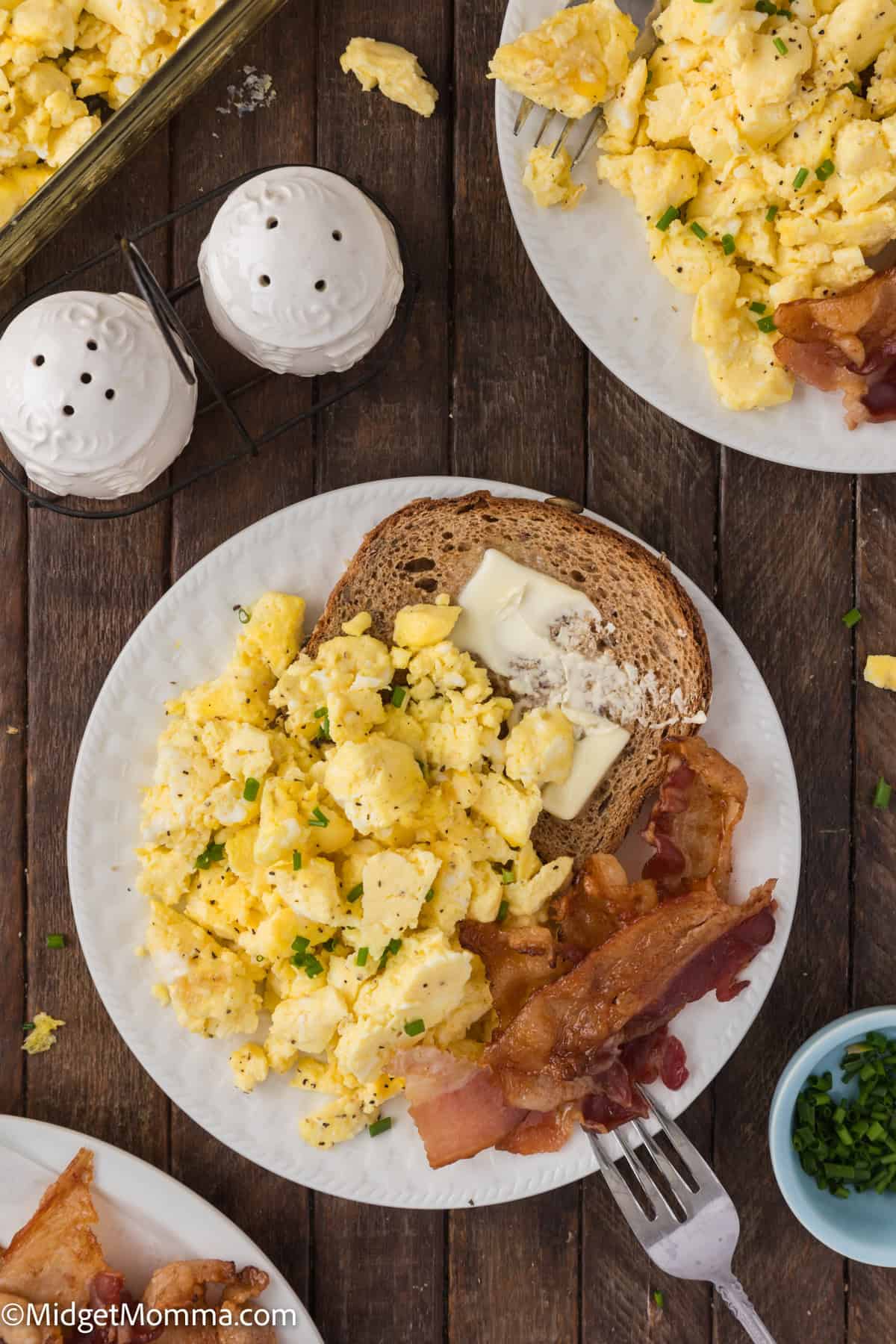 A plate with scrambled eggs, buttered toast, and bacon, garnished with herbs. Accompanied by salt and pepper shakers and a small bowl of chopped greens on a wooden table.