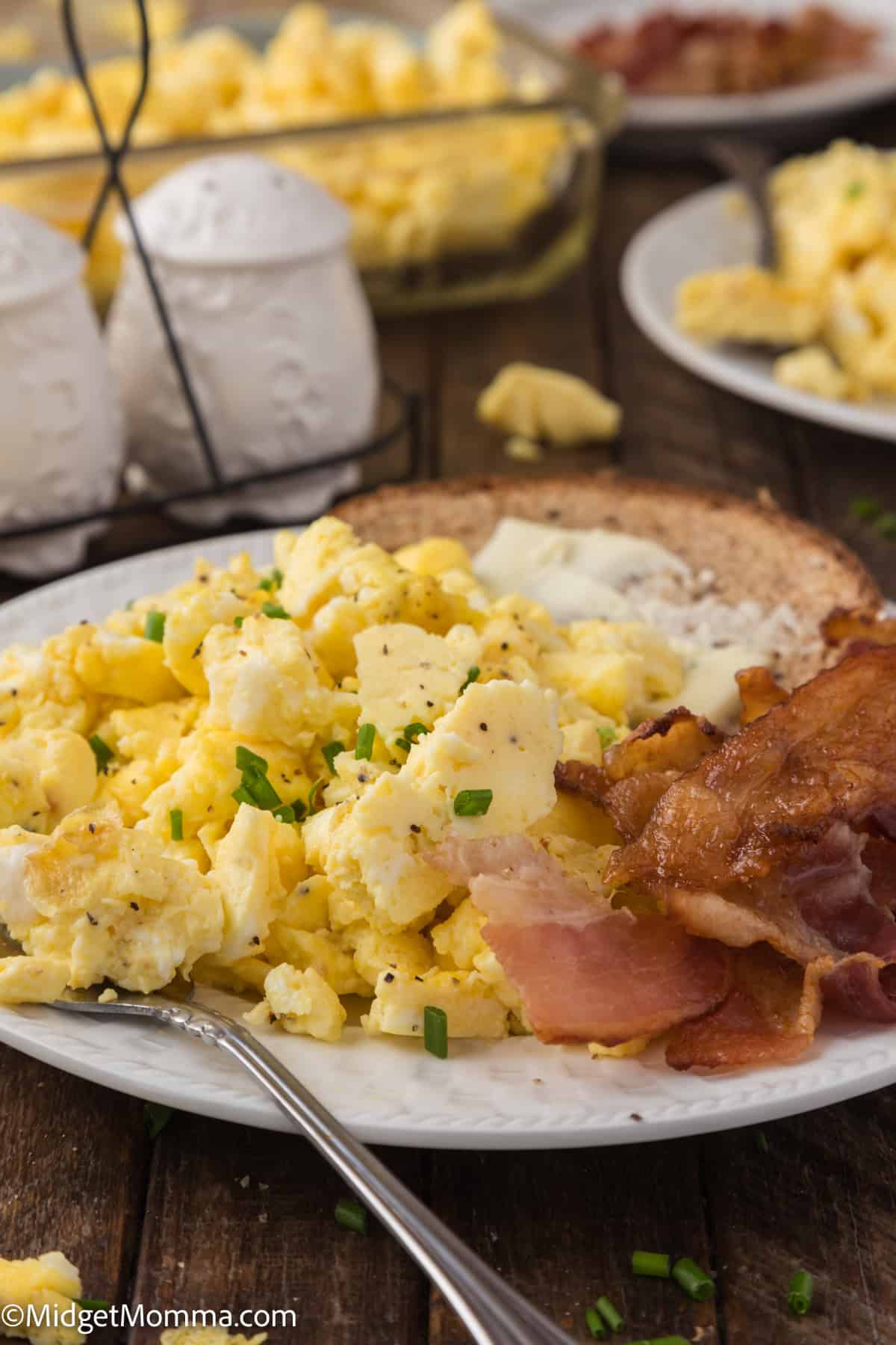 A plate of scrambled eggs garnished with chives, crispy bacon strips, and a slice of toast with butter. A fork rests on the plate.