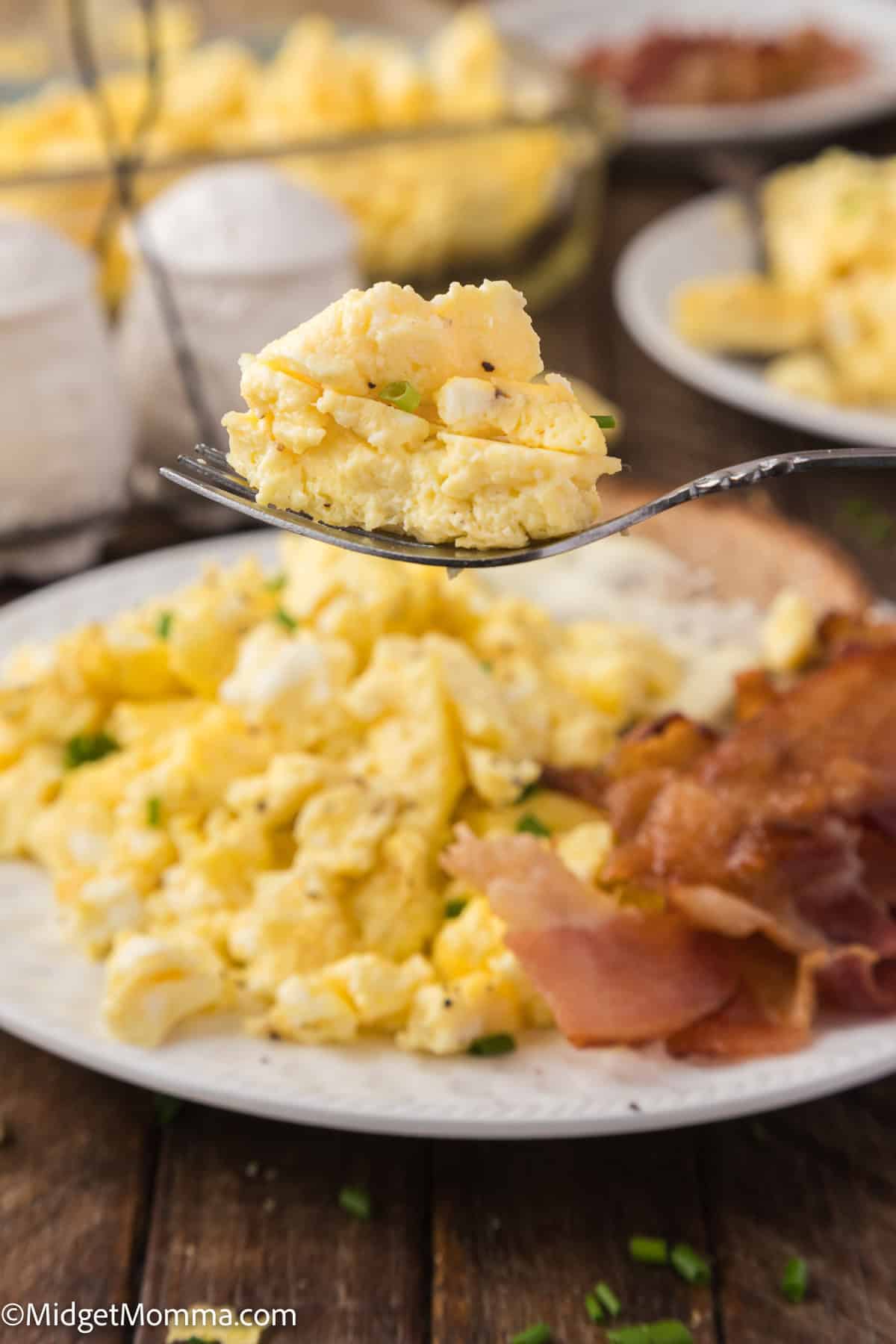 A fork with a bite of scrambled eggs is held above a plate with more scrambled eggs, bacon, and toast.