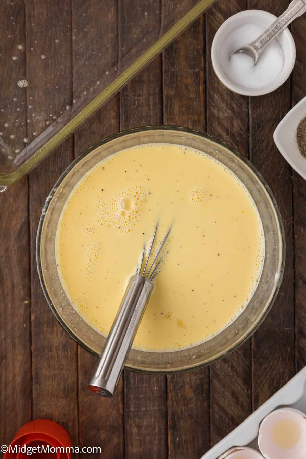 A bowl of scrambled egg mixture, with a whisk in it on a wooden table, surrounded by a baking dish, salt, pepper, and egg shells.