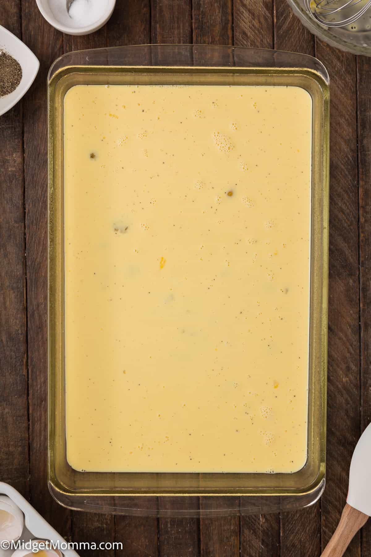 A rectangular glass baking dish filled with baked scrambled egg mixture mixture sits on a dark wooden surface. Nearby are small bowls and containers with various ingredients.