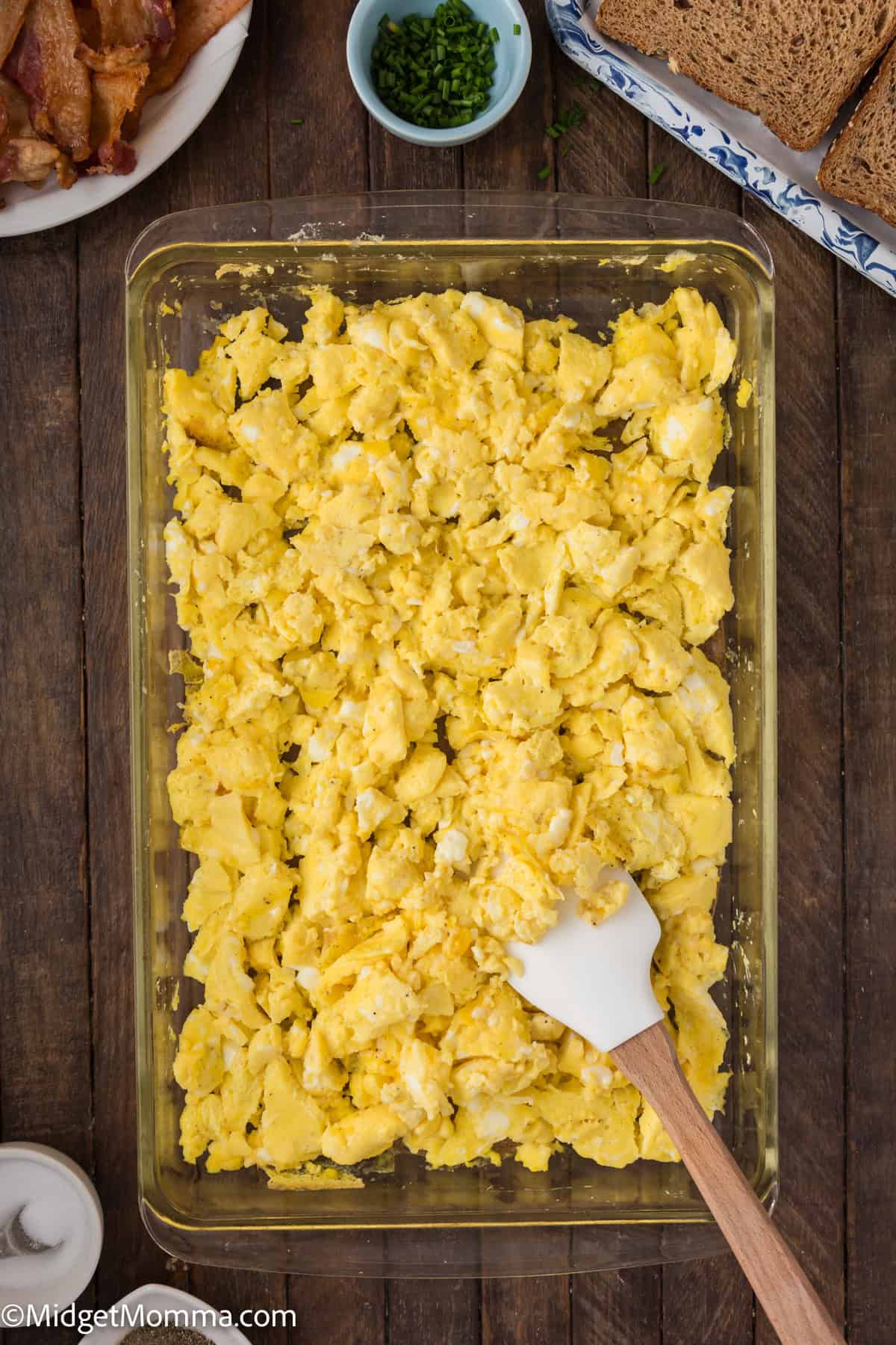 A clear glass baking dish filled with scrambled eggs. A white spatula with a wooden handle is placed on the right side of the dish. 