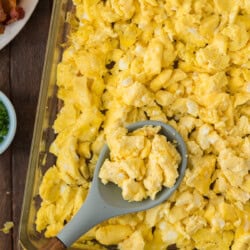 A baking dish filled with scrambled eggs; a serving spoon rests on top of the eggs. Part of a plate with bacon can be seen in the background.