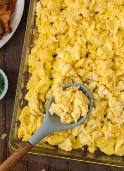 A baking dish filled with scrambled eggs; a serving spoon rests on top of the eggs. Part of a plate with bacon can be seen in the background.
