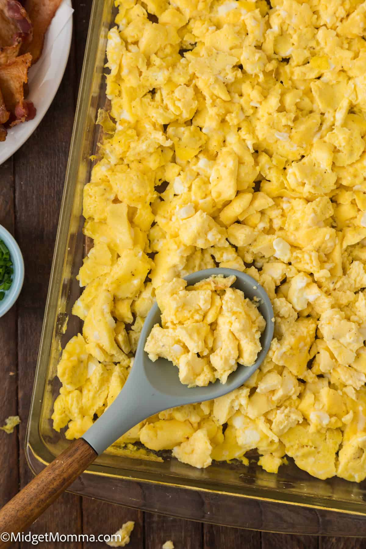 A baking dish filled with scrambled eggs; a serving spoon rests on top of the eggs. Part of a plate with bacon can be seen in the background.