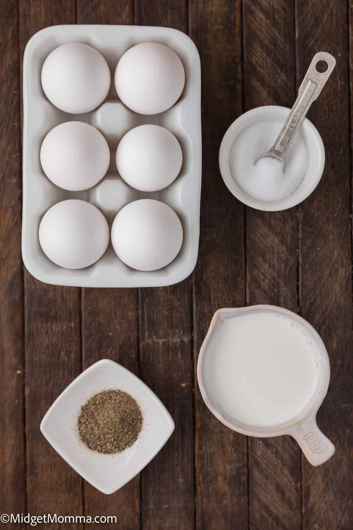 A flat-lay view of six eggs in a carton, a bowl of salt with a measuring spoon, a small bowl of black pepper, and a measuring cup of milk on a wooden surface.