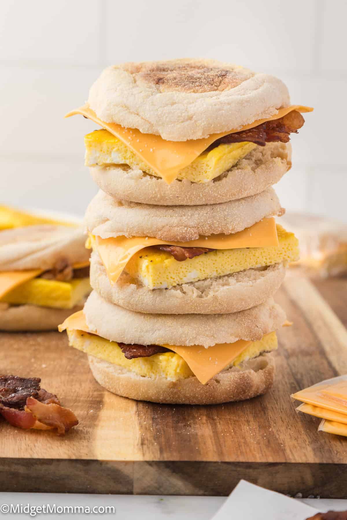 A stack of three breakfast sandwiches with English muffins, scrambled egg, bacon, and cheese on a wooden surface.