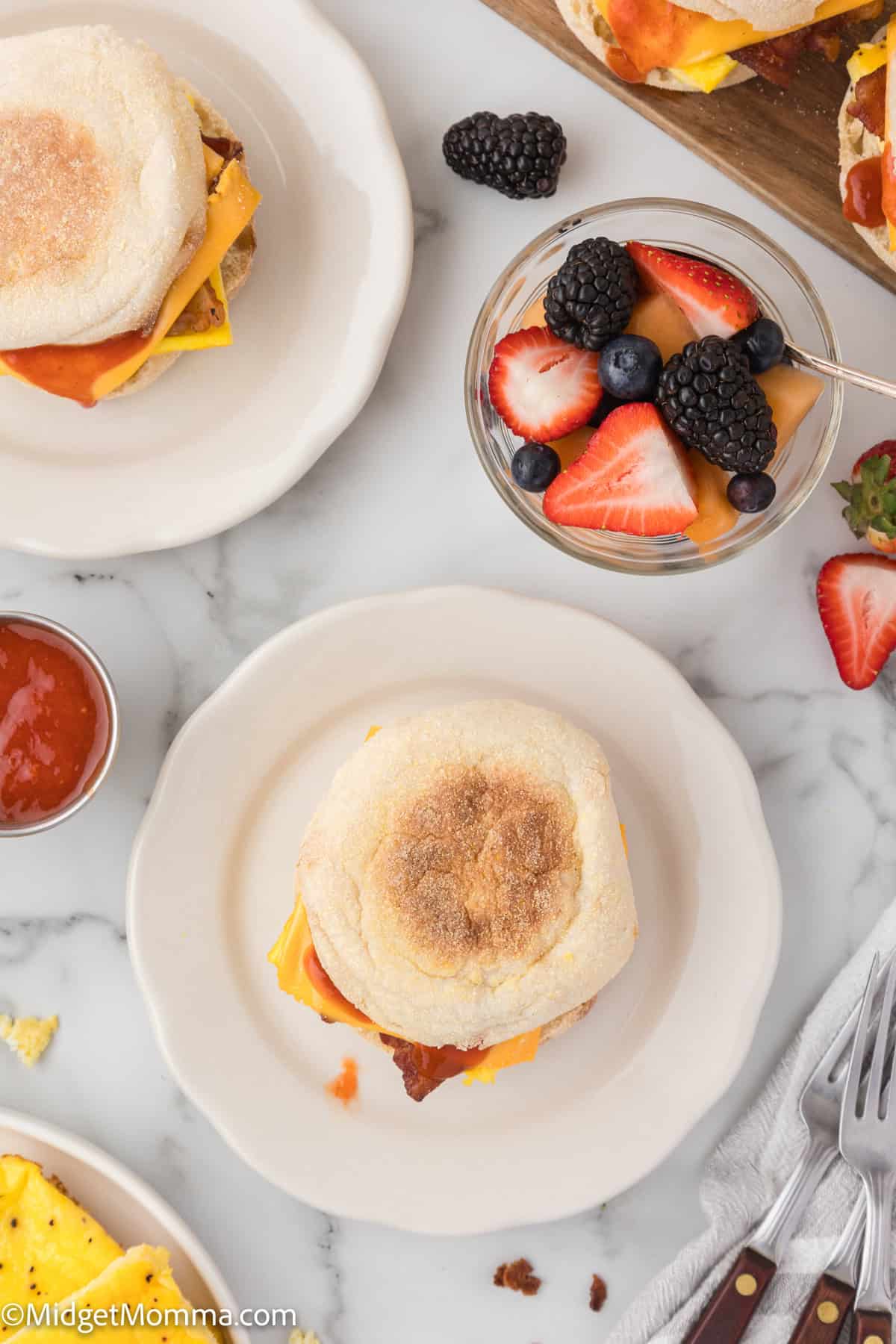 Two plates with breakfast sandwiches and a bowl of mixed berries on a white marble surface. Ketchup is in a small dish nearby. Forks and scattered fruit pieces are visible. 