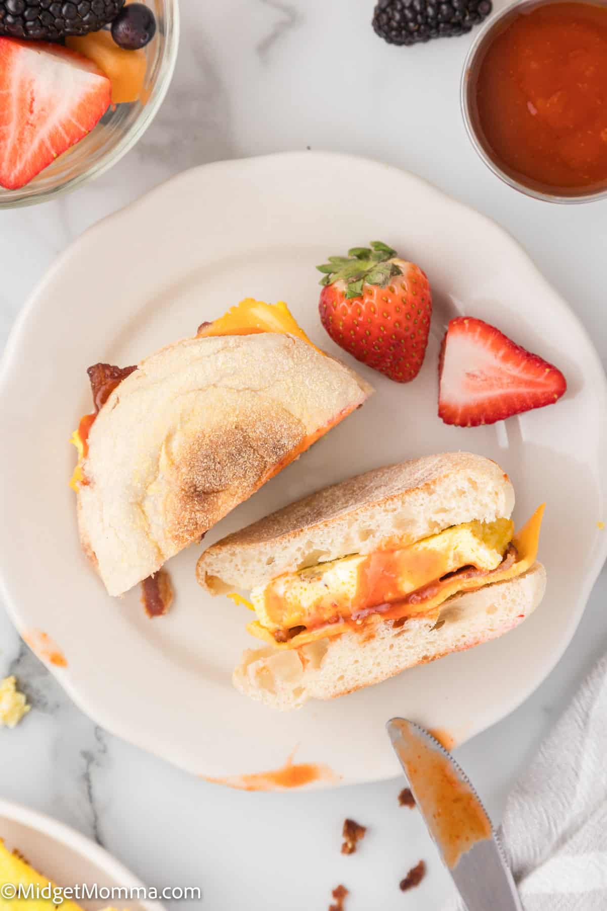 A breakfast sandwich with egg, cheese, and bacon on a white plate, accompanied by sliced strawberries, a bowl of fruit, and a small dish of sauce on a marble countertop.