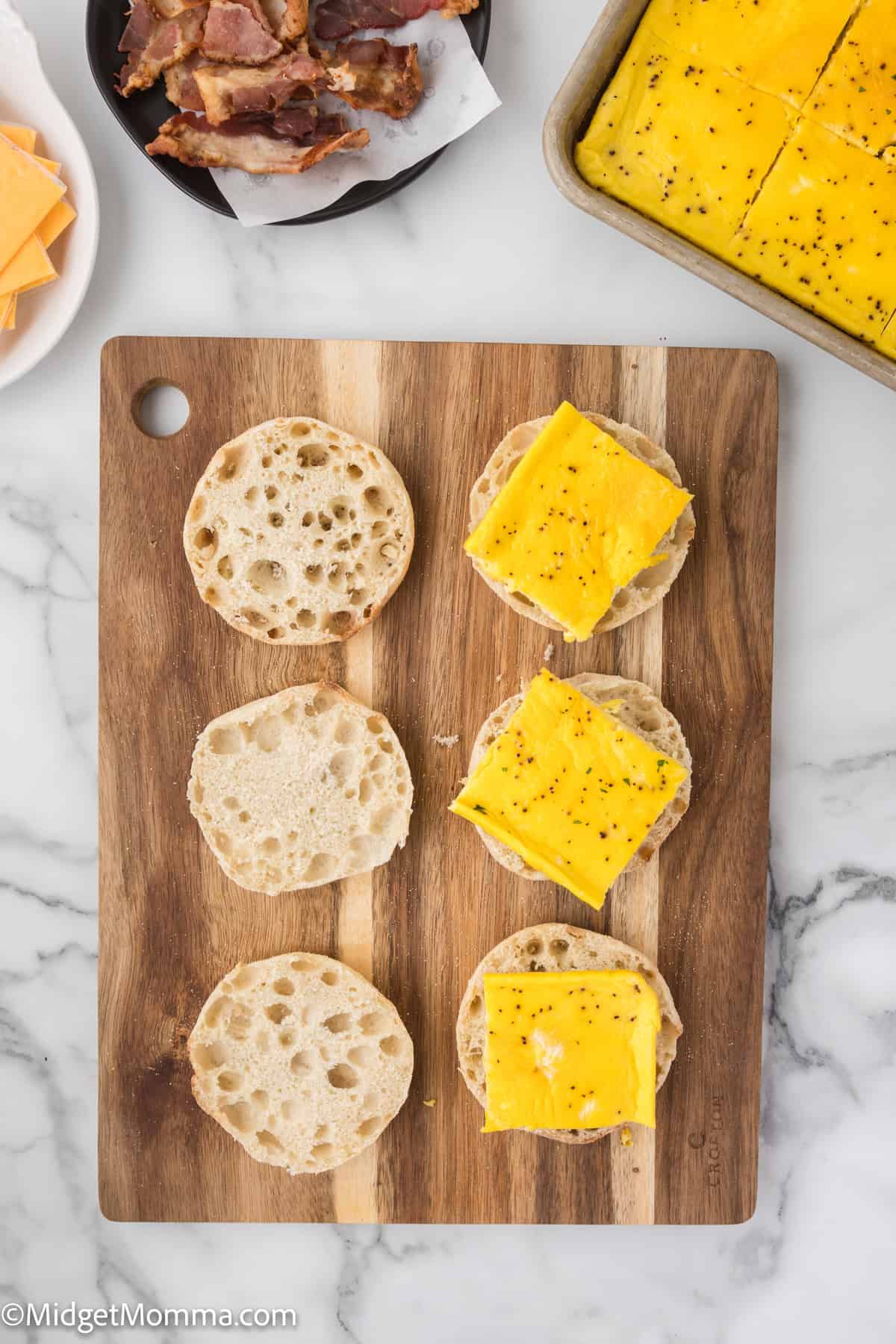 A wooden board with three split English muffins, two topped with cheese and black pepper. Nearby are sliced cheese, bacon, and a baking dish with cheese-topped slices.