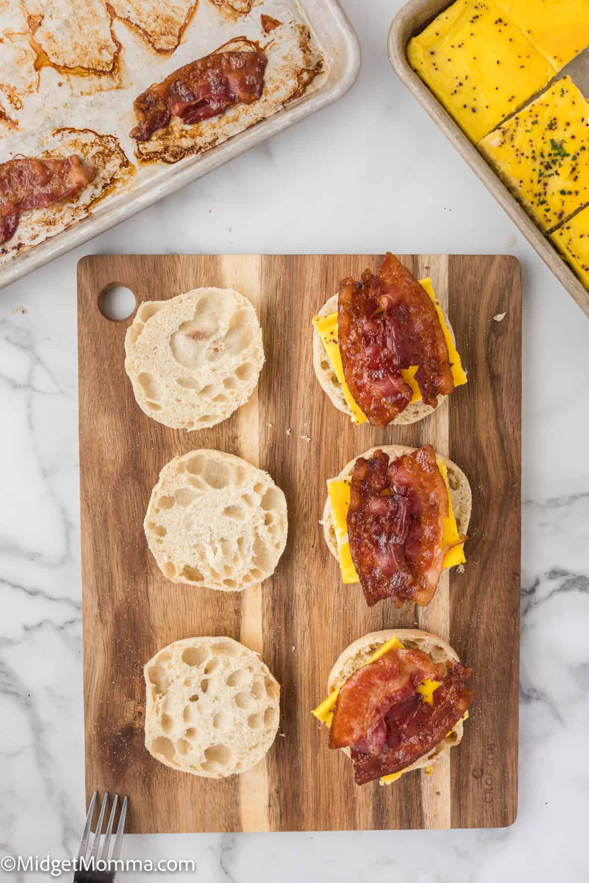 A wooden board with open English muffins, each topped with a slice of cheddar cheese and bacon. Trays with more bacon and cheese are visible in the background.