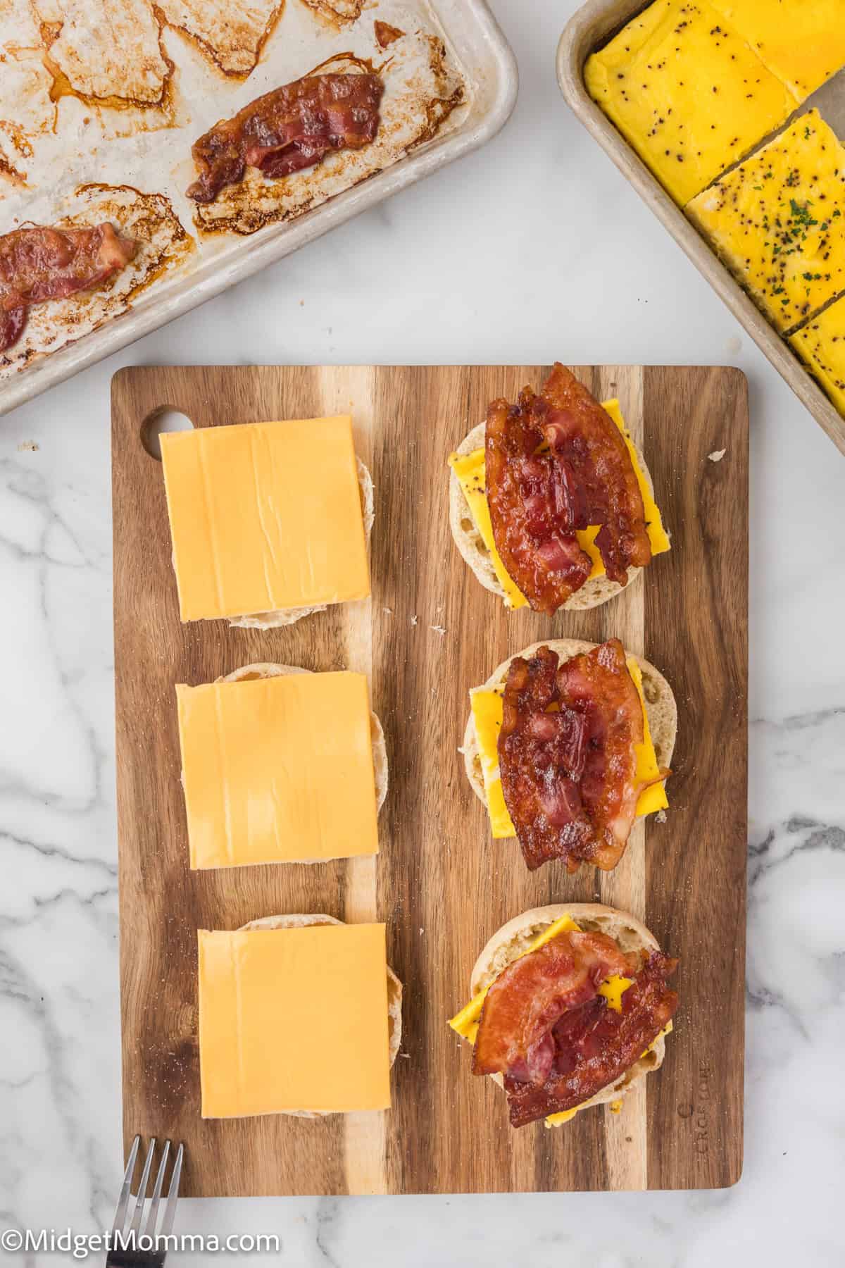 Wooden board with four English muffin halves, each topped with cheese slices and bacon. Nearby, a tray with cooked bacon and a pan containing sliced yellow cheese are visible.