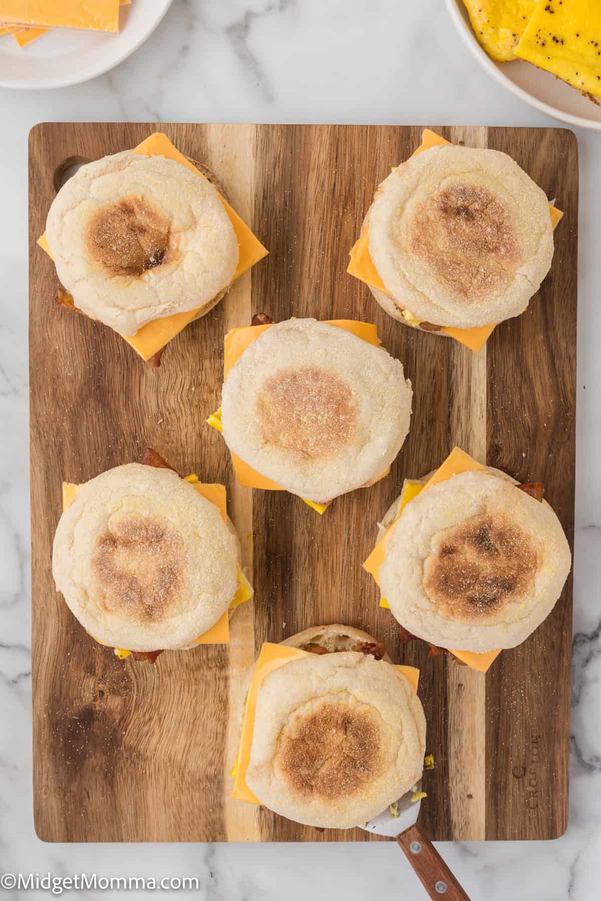 A wooden board with six English muffins, each filled with melted cheese, displayed on a marble surface.