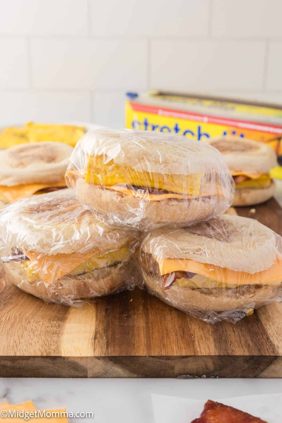 Three wrapped breakfast sandwiches sit on a wooden cutting board. In the background, a box of plastic wrap and other food items are visible.