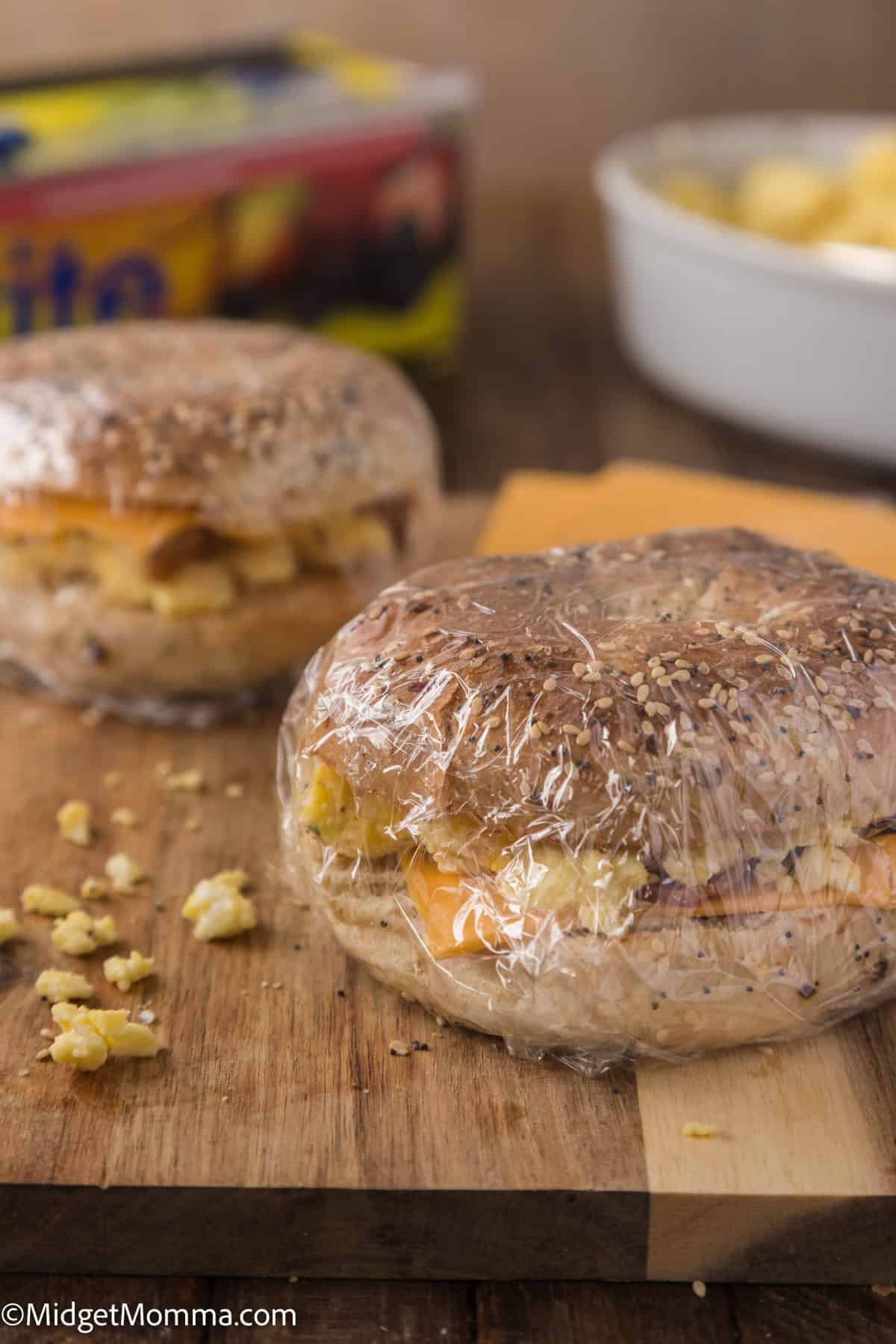 Two bagel sandwiches with visible fillings wrapped in plastic wrap are placed on a wooden surface. A container with blurred text and another dish with food are visible in the background.