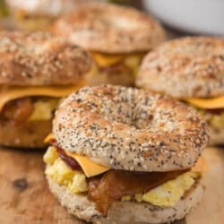 Four loaded bagel sandwiches, each filled with scrambled eggs, crispy bacon, and cheddar cheese, displayed on a wooden surface. A bowl of potato chips is blurred in the background.