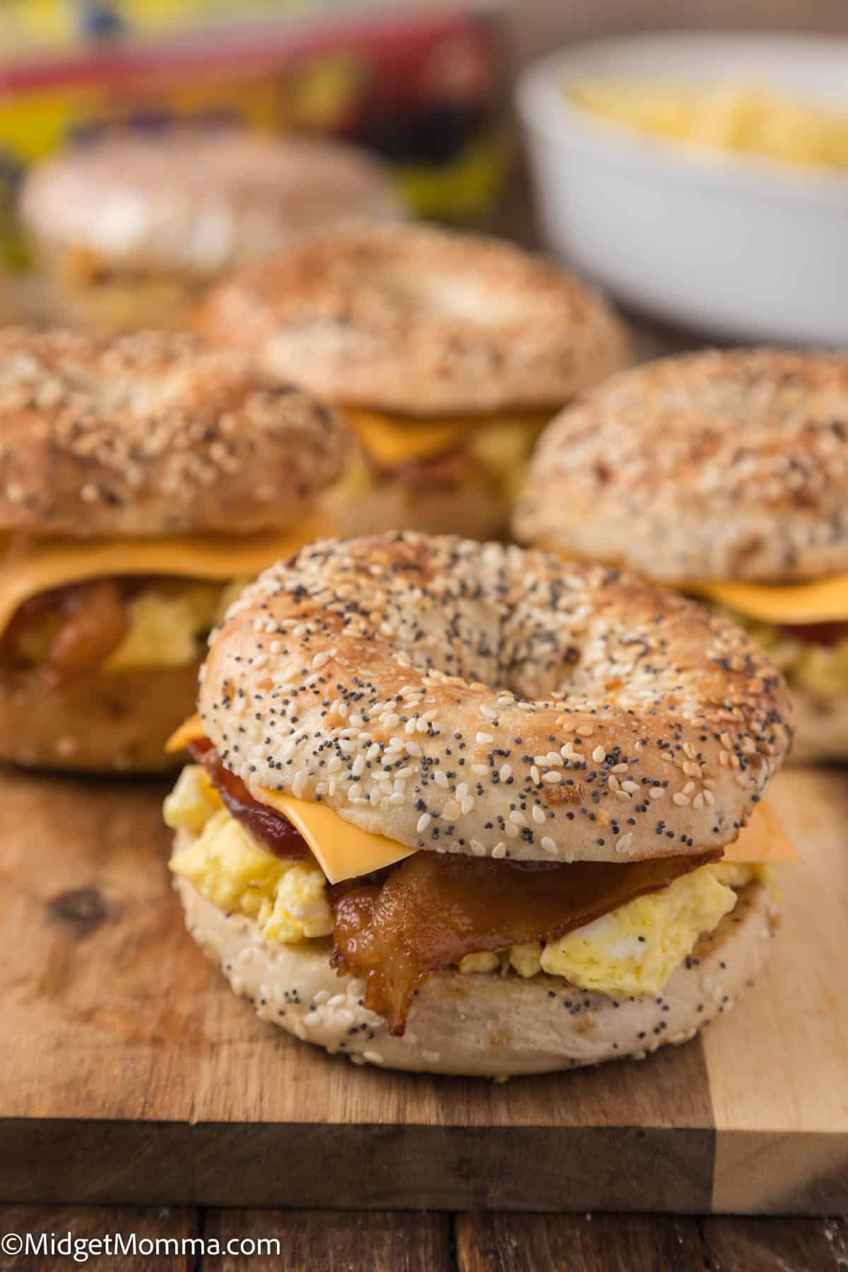 Four loaded bagel sandwiches, each filled with scrambled eggs, crispy bacon, and cheddar cheese, displayed on a wooden surface. A bowl of potato chips is blurred in the background.