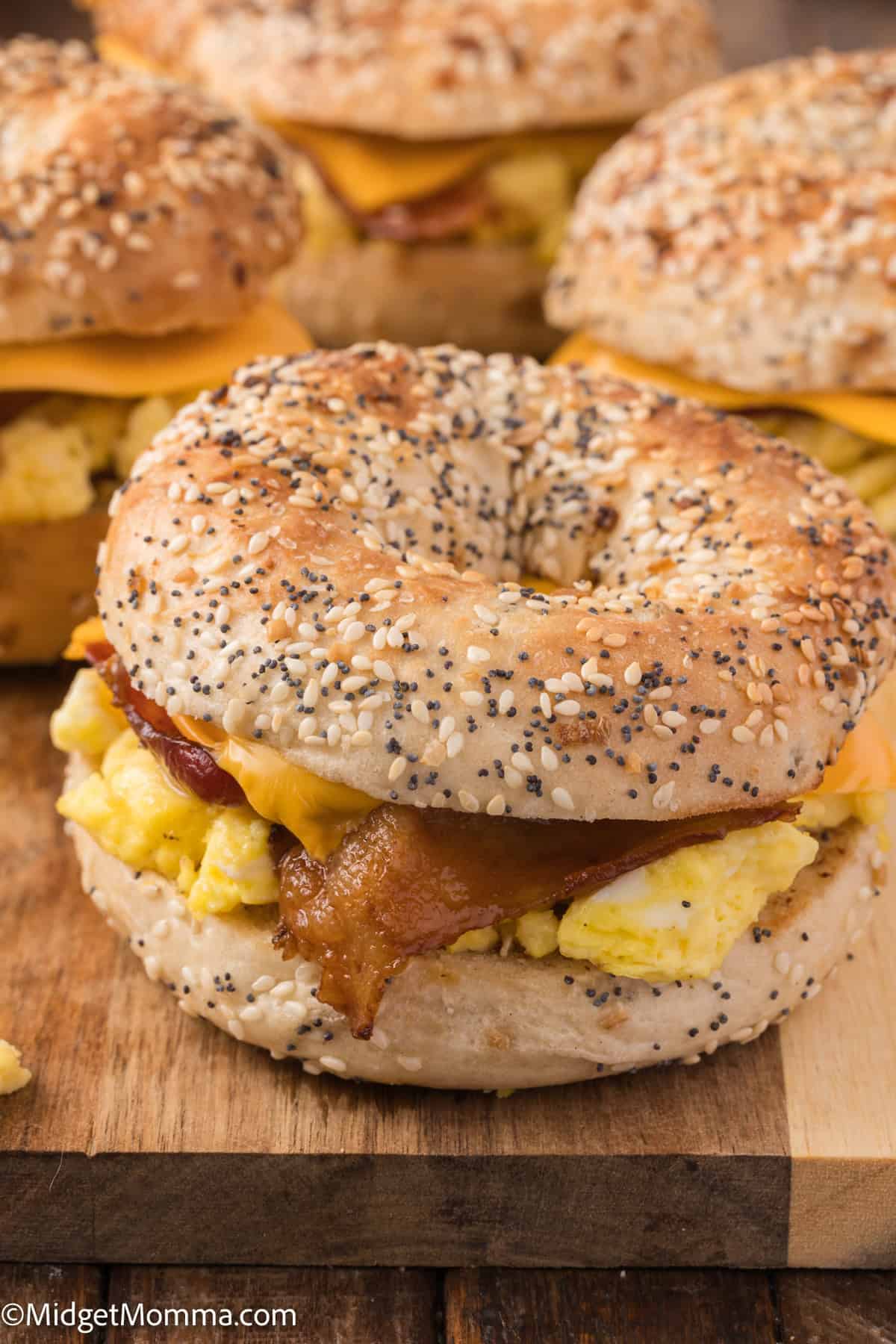 A close-up of a breakfast sandwich made with a seeded bagel, scrambled eggs, bacon, and cheese, placed on a wooden surface with similar sandwiches in the background.