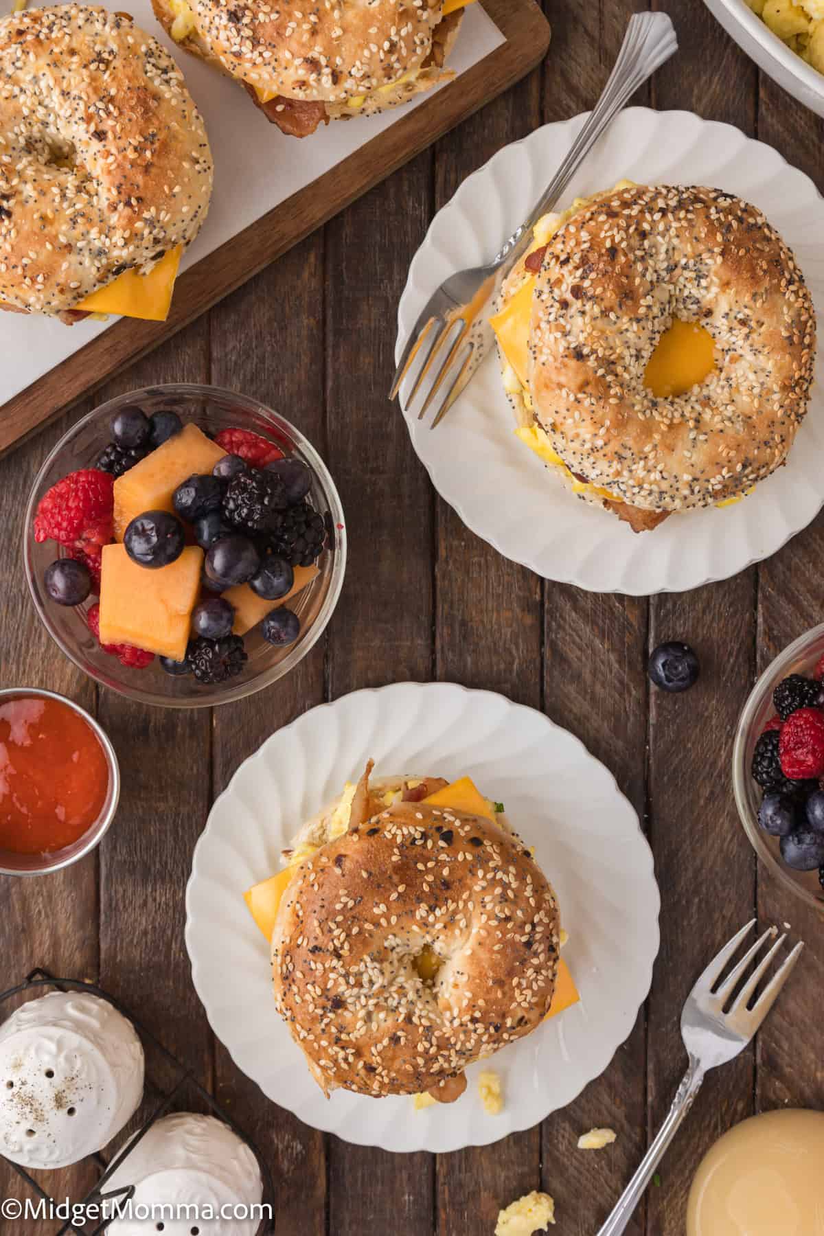 Top-down view of a breakfast setting featuring two sesame bagel sandwiches with cheese on plates, bowls of mixed fruit, a bowl of jam, and a cup of coffee on a wooden table.