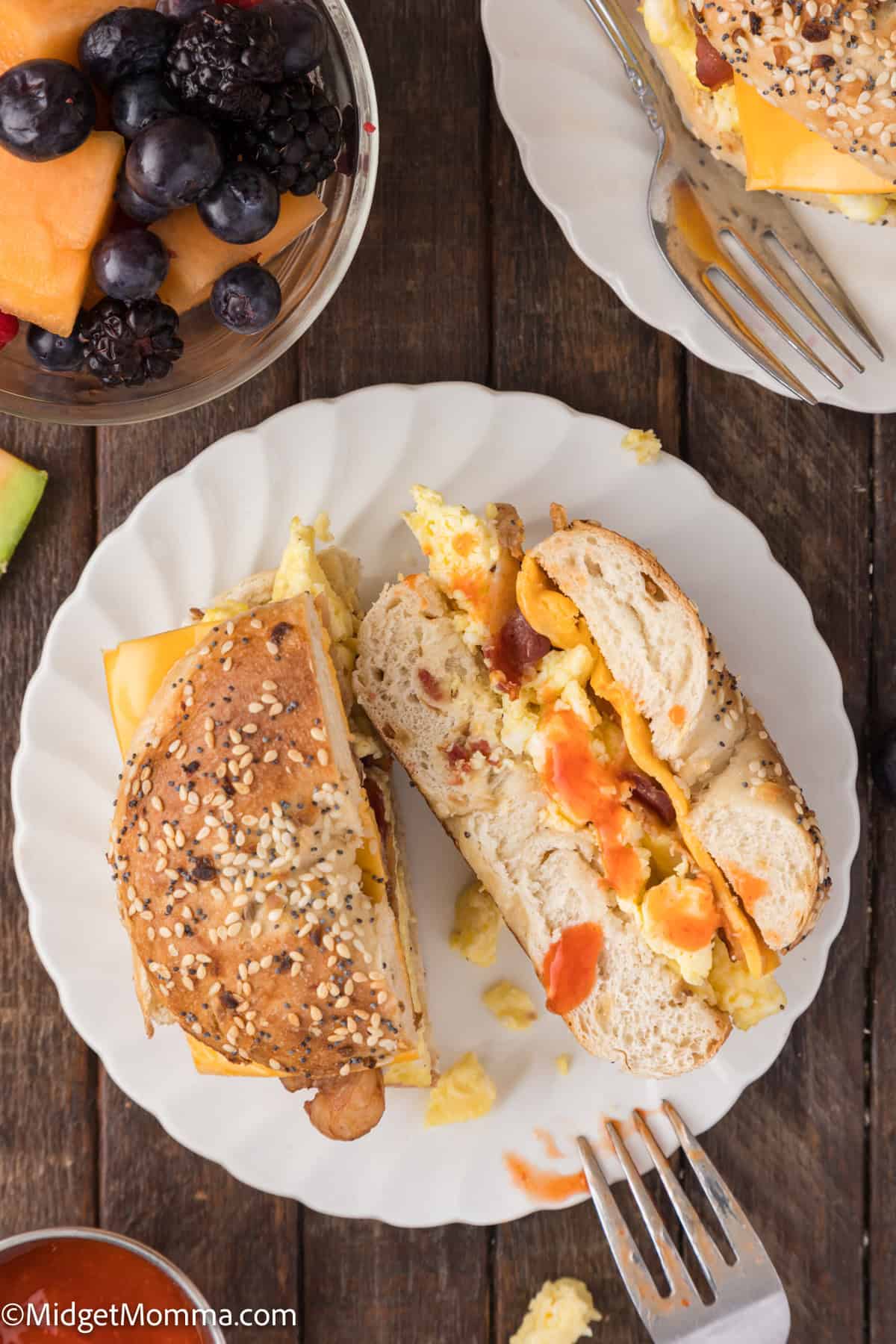 A bagel sandwich with scrambled eggs, cheese, and bacon is sliced in half on a white plate. A fork is beside the sandwich. Fresh fruit is in a bowl nearby.