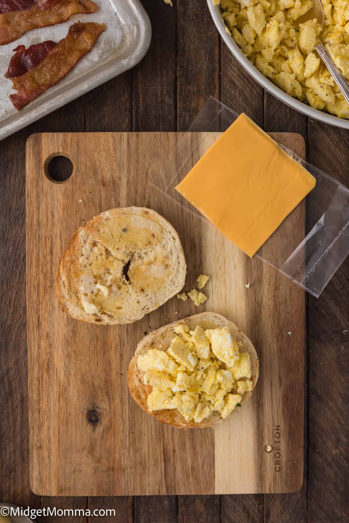 A wooden cutting board with a sliced bagel topped with scrambled eggs, a slice of cheese, and a plate of bacon in the background.