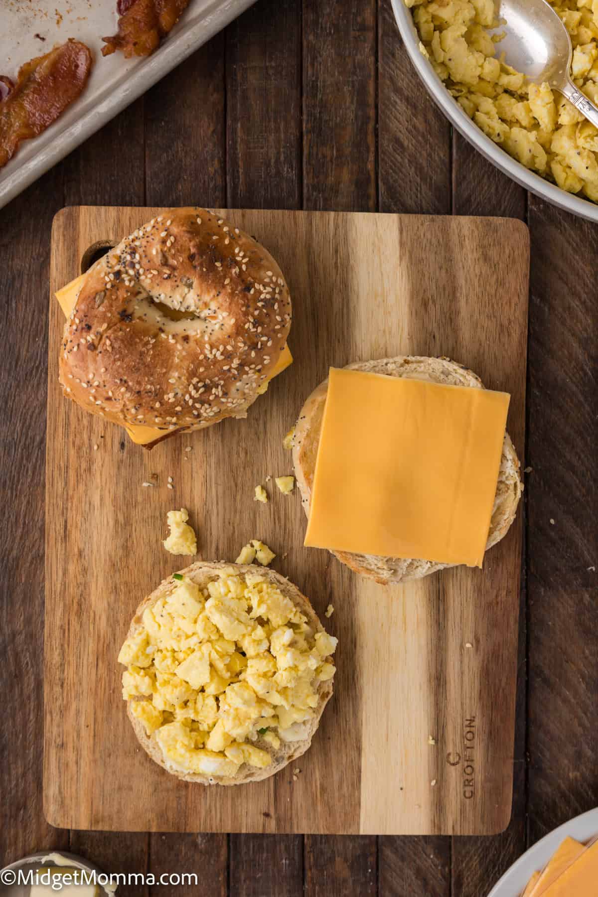 A wooden cutting board with a bagel sandwich being assembled, showing a bagel half with scrambled eggs and another with a slice of cheese. Nearby, a tray with bacon and a bowl of scrambled eggs.