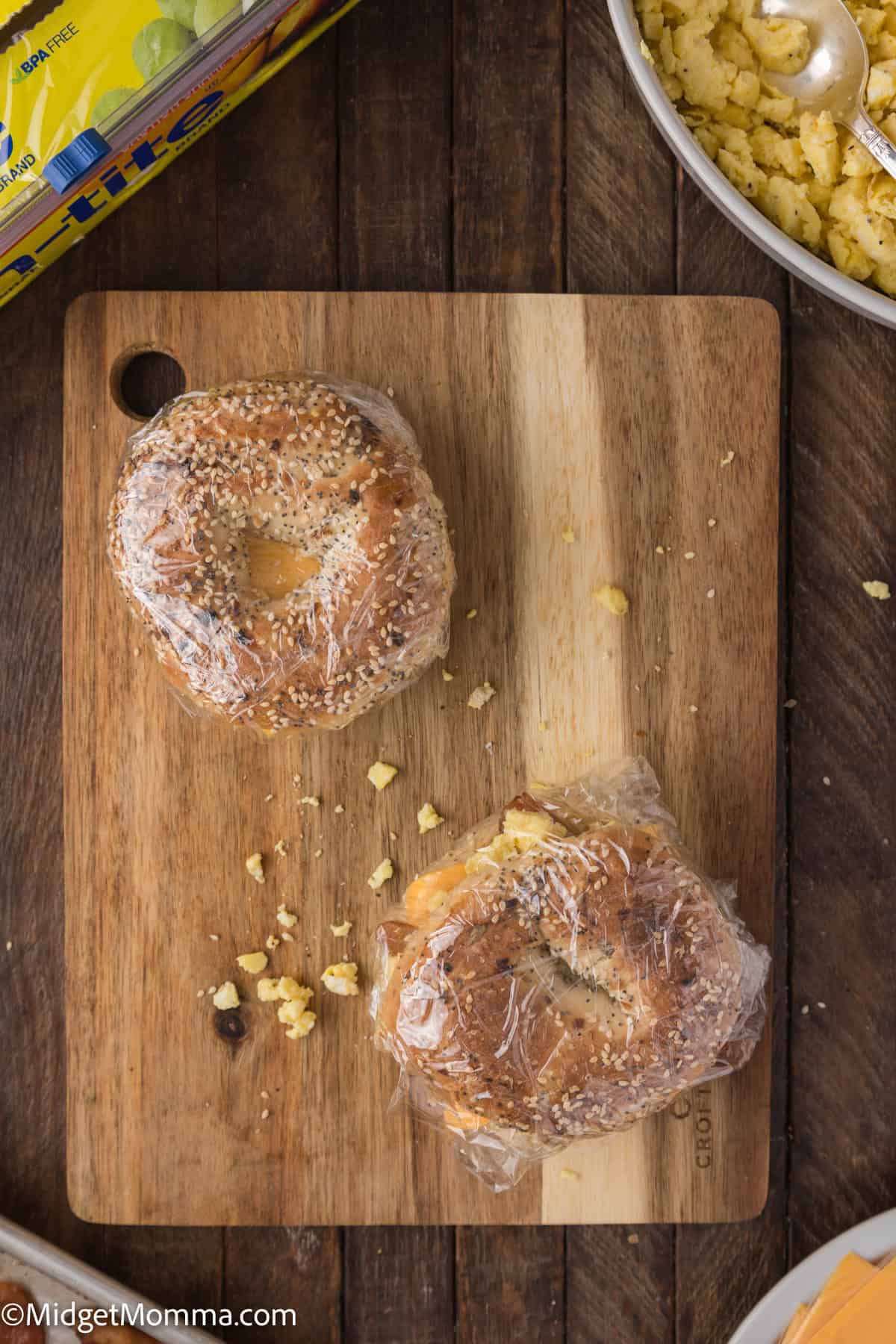 Two bagels wrapped in plastic wrap sit on a wooden cutting board surrounded by crumbs and partially out-of-frame kitchen items.