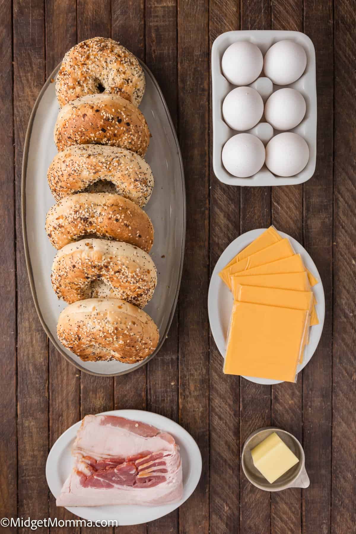 A wooden table with bagels, a carton of eggs, slices of cheese, strips of raw bacon, and a small dish of butter.