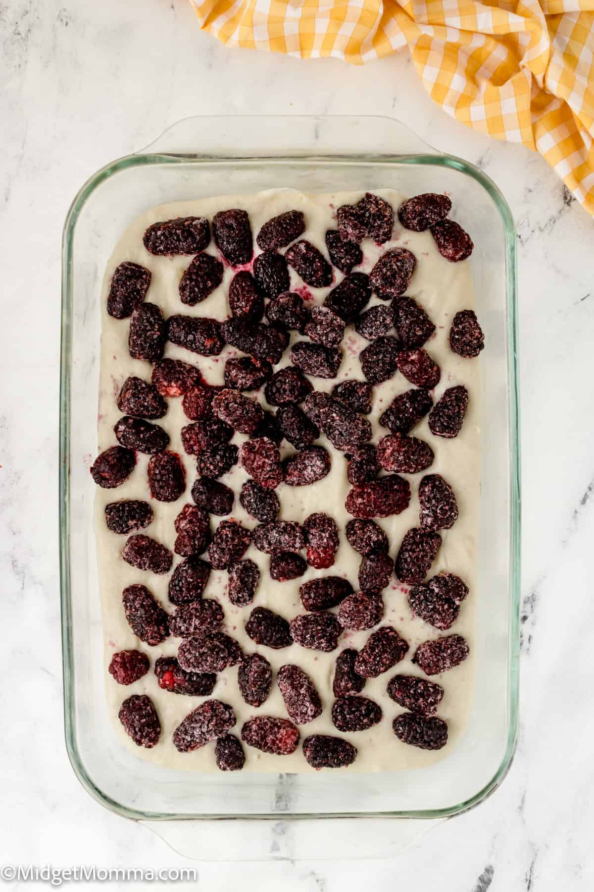 A glass baking dish containing batter with a layer of blackberries on top, placed on a marble surface. A yellow and white checkered cloth is partially visible in the corner.