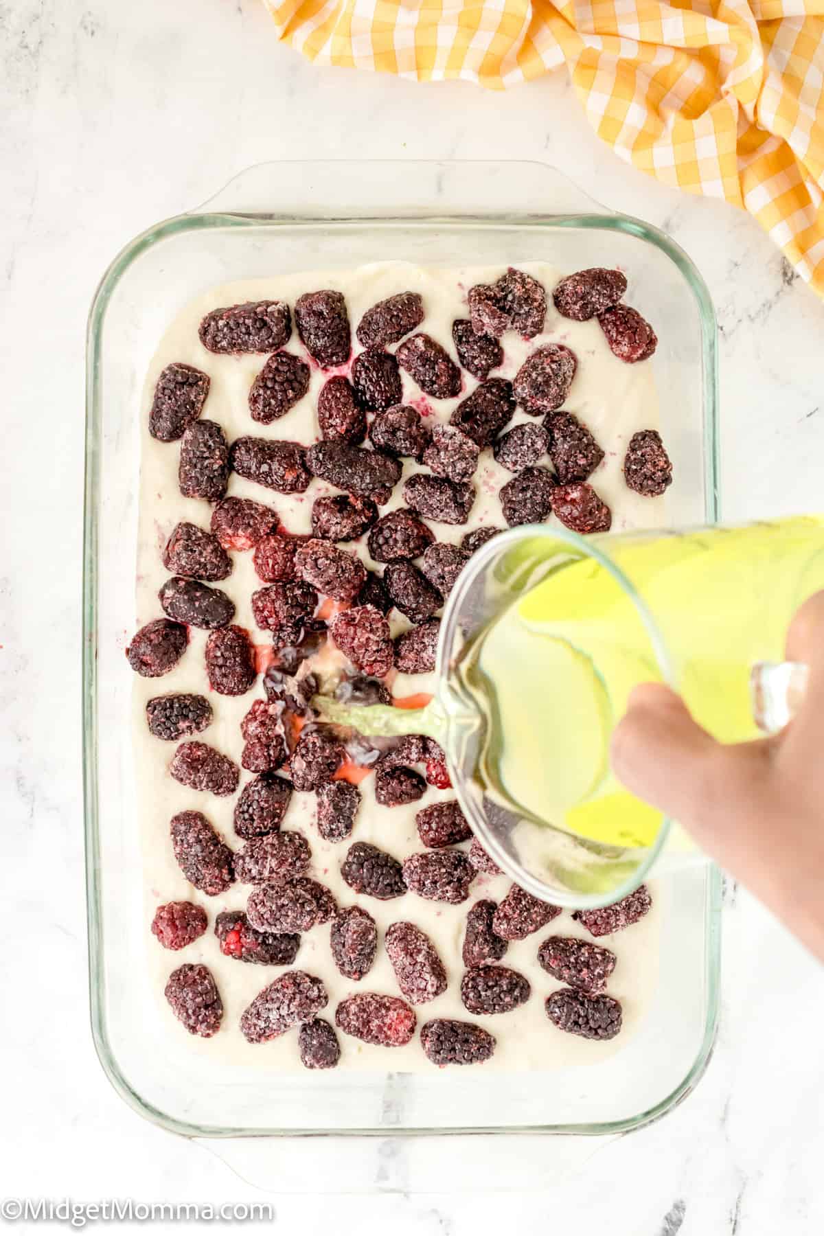 A hand pours liquid from a measuring cup into a glass baking dish filled with a creamy mixture and topped with frozen raspberries. A yellow checkered cloth is partially visible on the side.